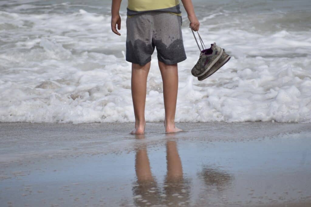 Photo of Rocket Kid's legs with wet shorts, standing on water's edge, holding his sneakers.