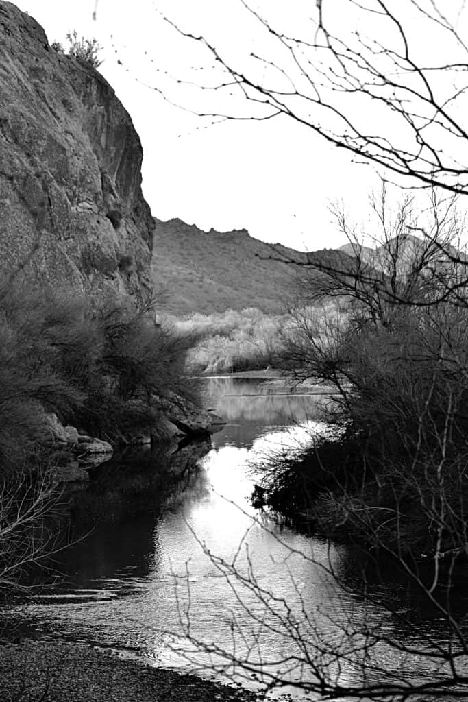 black and white photo with the river in the center, bushes and trees to the right, a rock face to the left, and mountains in the background